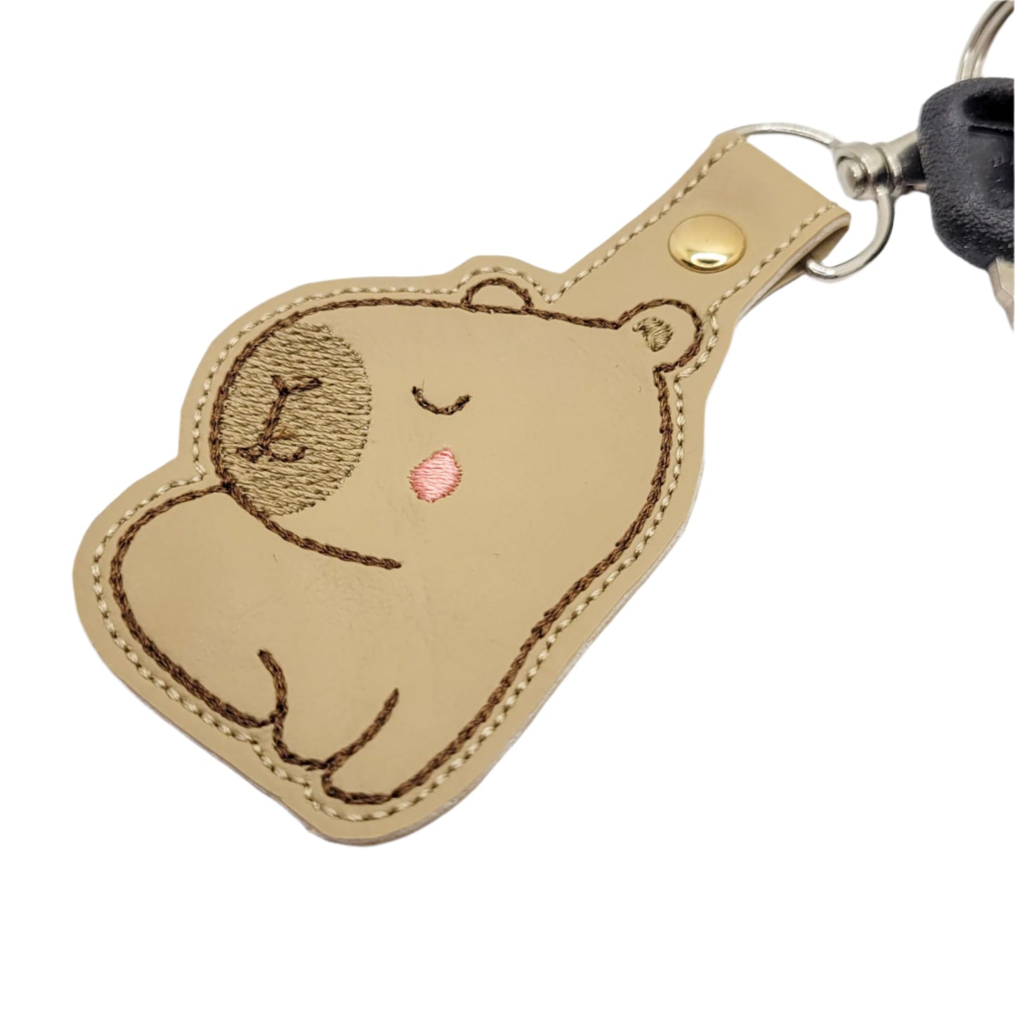 Embroidered vinyl keychain shaped like a capybara, with eyes closed and a pink cheek, attached to a set of keys