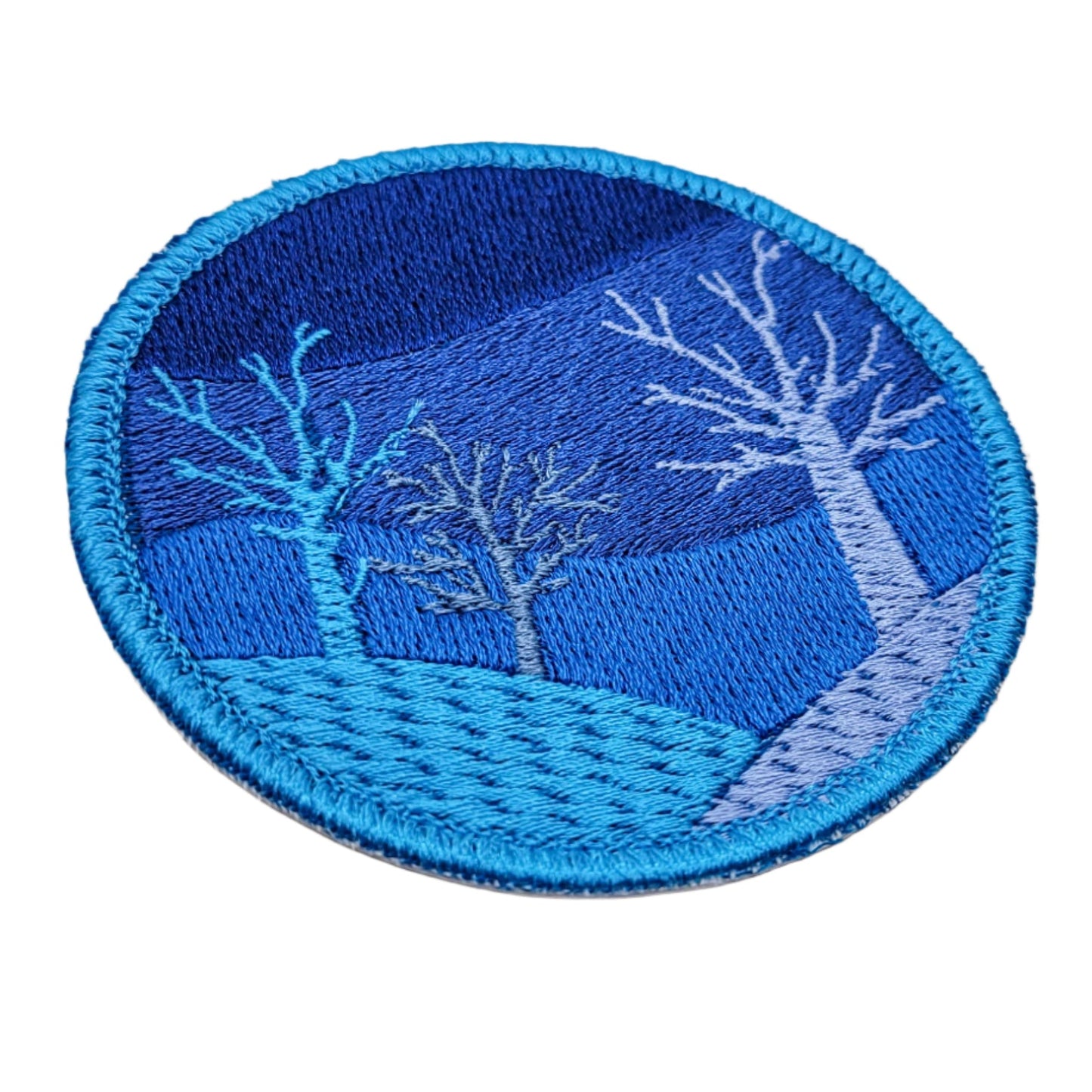 Circular patch with embroidered design of trees on a blue background, placed on a white background.