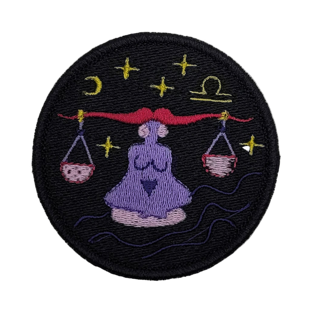 Embroidered patch of a person in a meditative pose with scales and zodiac symbols on a black background.