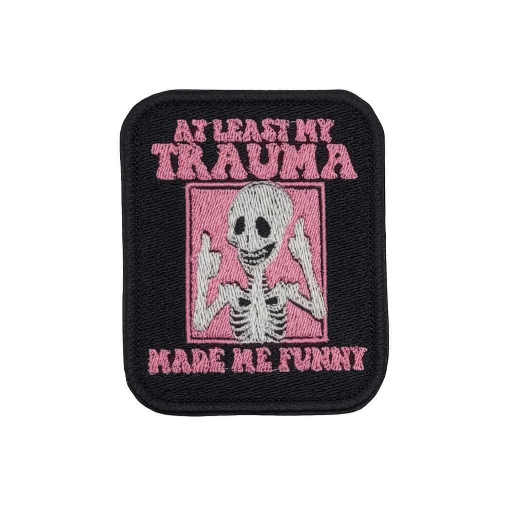 A rectangular embroidered patch with a pink background featuring a skeleton giving a thumbs up sign with the text 'At Least My Trauma Made Me Funny'.