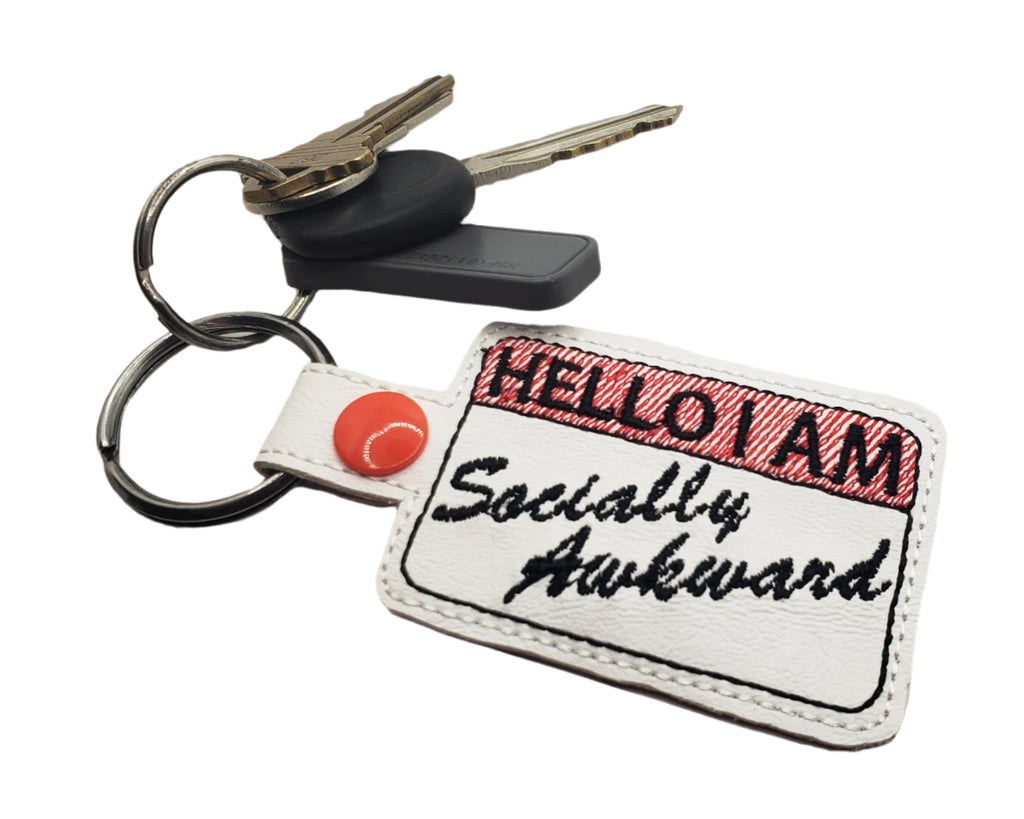 Embroidered vinyl keychain with a red-and-white name tag design that reads “Hello I Am Socially Awkward,” shown attached to a set of keys.